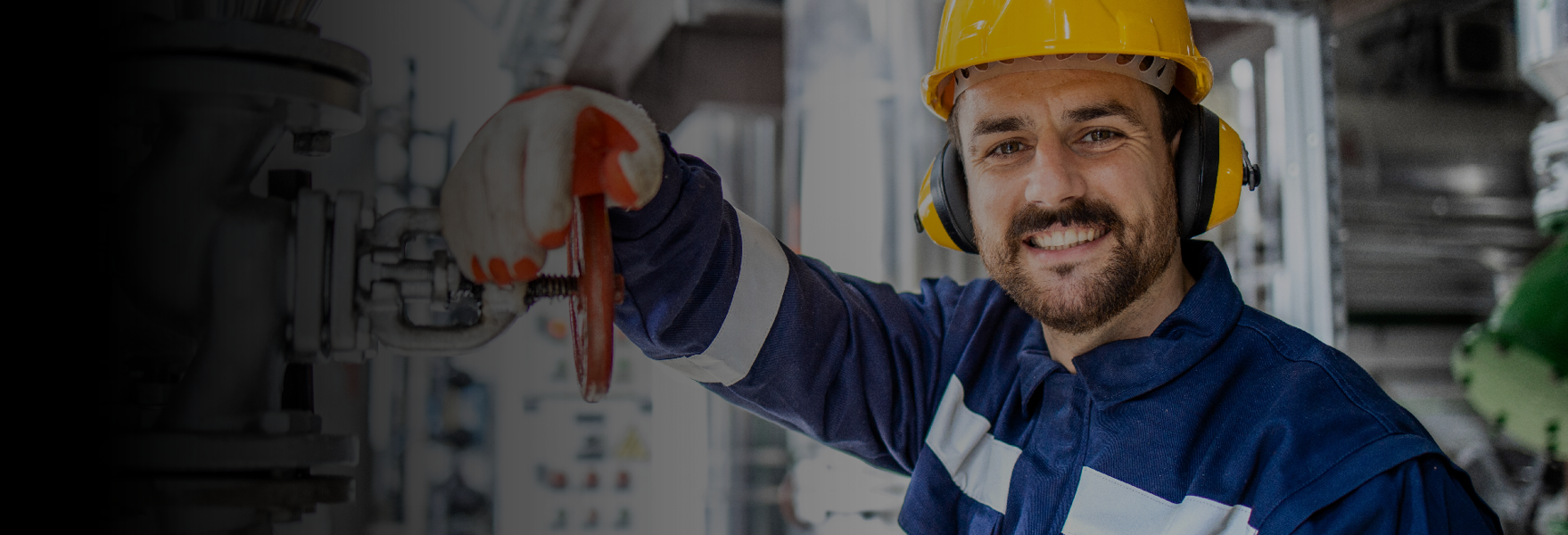 Technician in a blue work shirt and yellow hard hat and ear muffs smiling at the camera in front of an industrial background